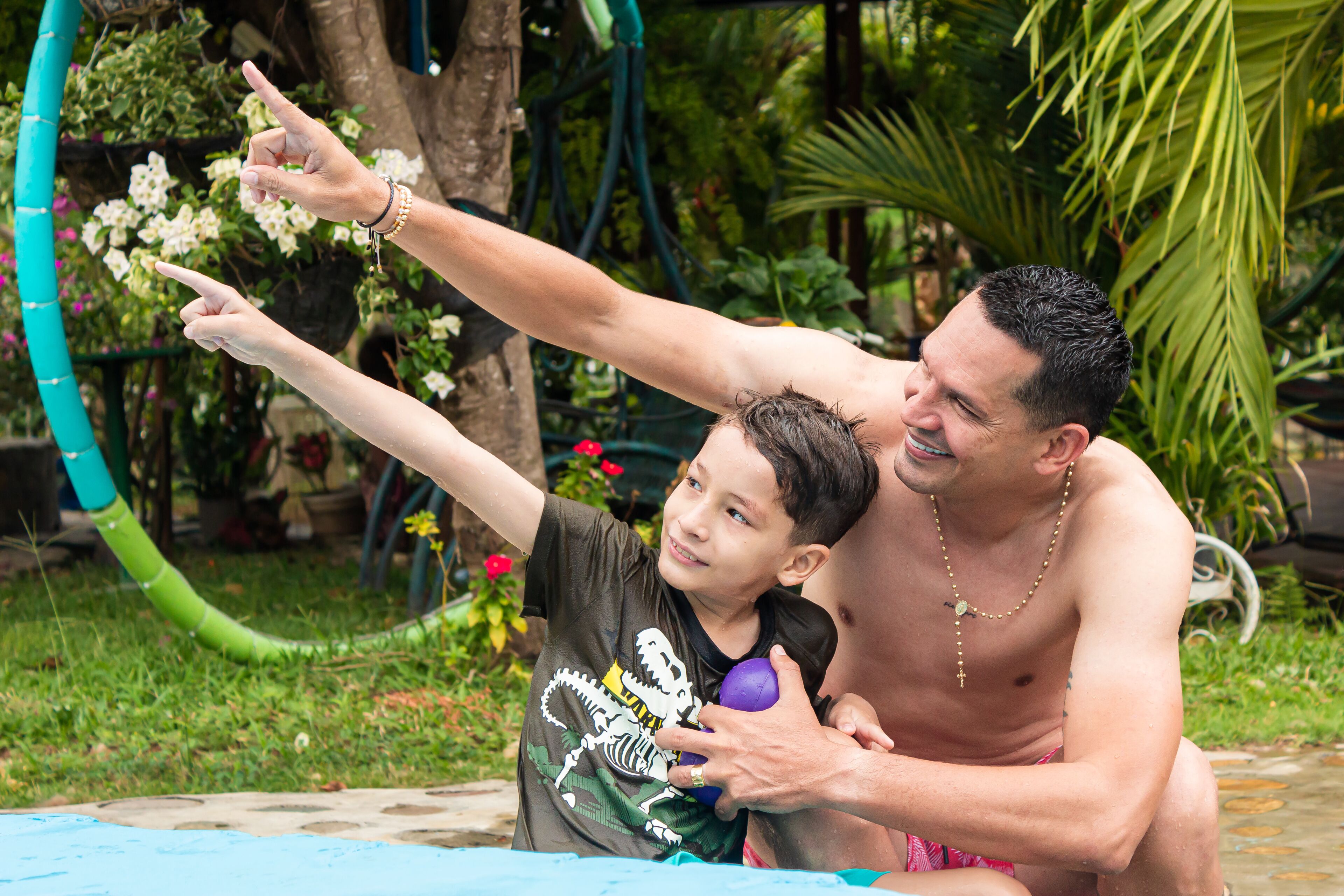 Un hombre y un niño junto a una piscina, con follaje y flores al fondo.