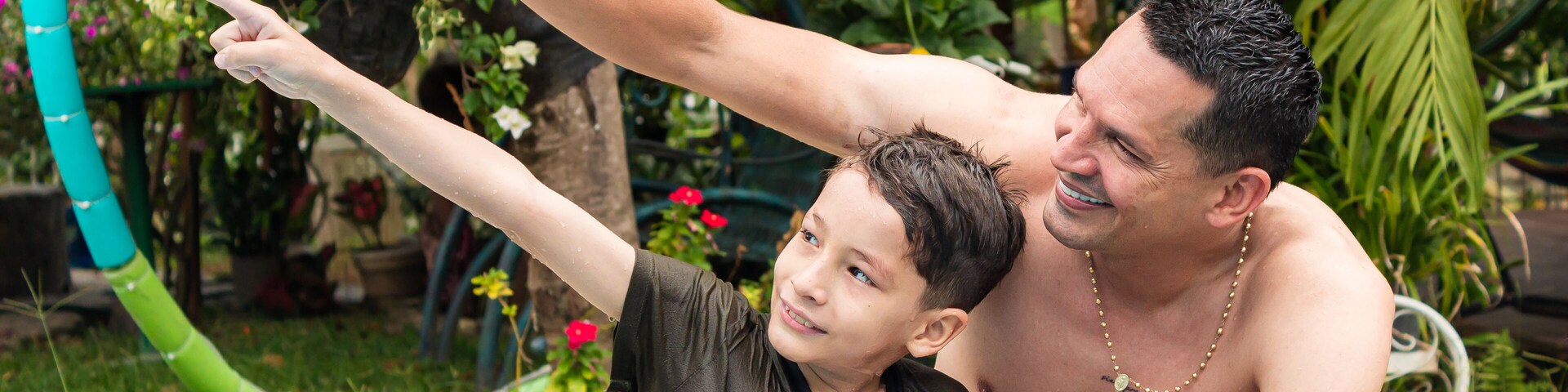 Un hombre y un niño junto a una piscina, con follaje y flores al fondo.