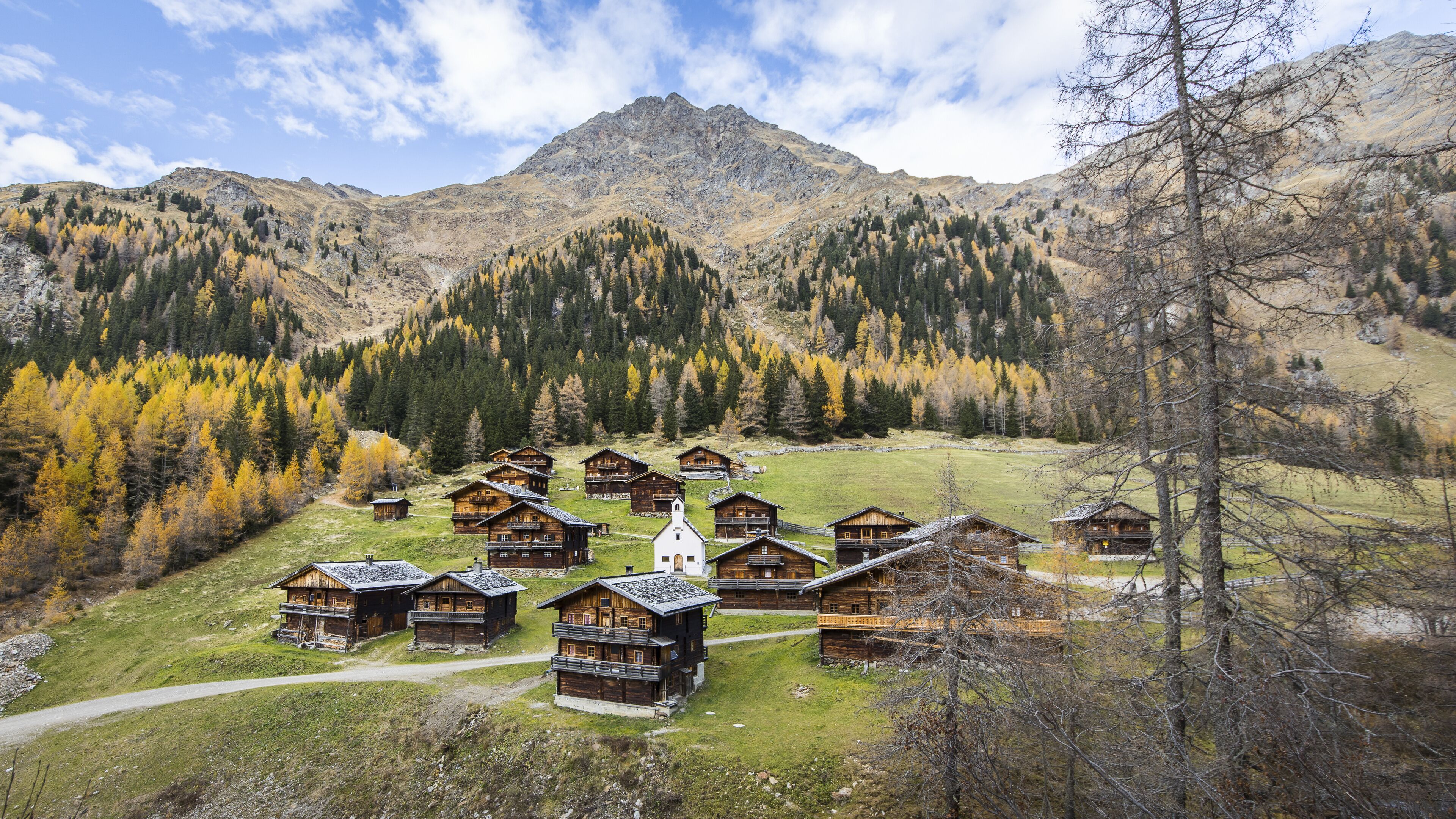 View of rustic wooden chalets nestled in a valley, surrounded by golden-hued autumn trees and towering mountains under a sky with scattered clouds, Innervillgraten, Tyrol, Austria.