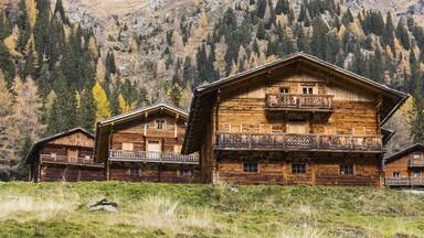 View of rustic wooden chalets nestle on a grassy hill amidst a backdrop of autumnal trees climbing towards the rugged mountain peaks, Innervillgraten, Tyrol, Austria.
