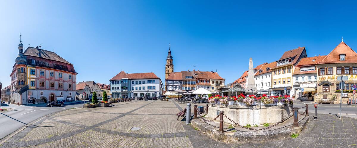 A cobblestone square with historic buildings, a church tower, and a fountain with flowers.