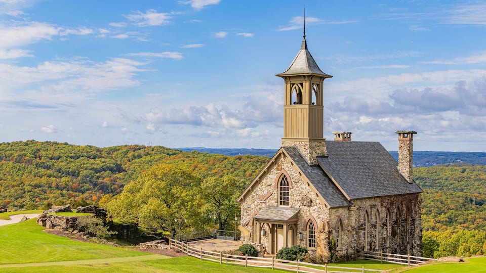 A stone church with a bell tower, surrounded by a lush green landscape and a forested hill in the background.