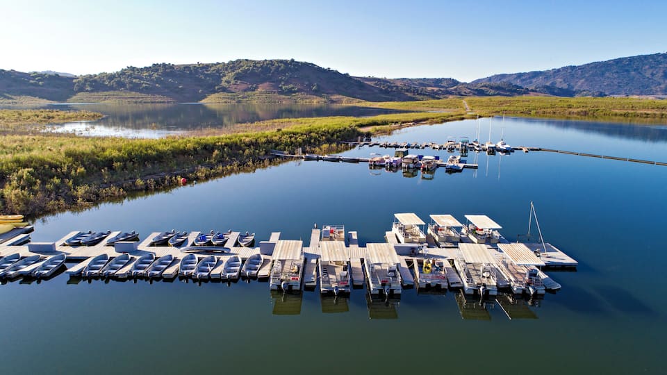 A marina with boats docked on wooden piers, surrounded by calm water and green hills in the background.
