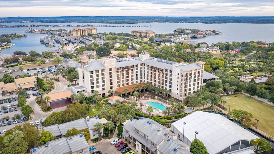 A large hotel with a swimming pool surrounded by lush greenery and a lake in the background.