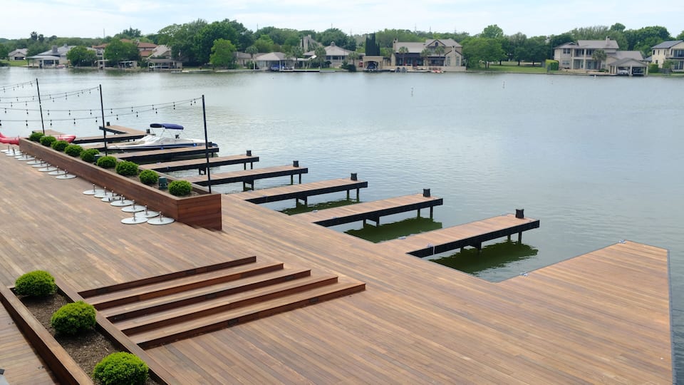 A wooden deck with benches and small trees, overlooking a calm lake with houses in the background.