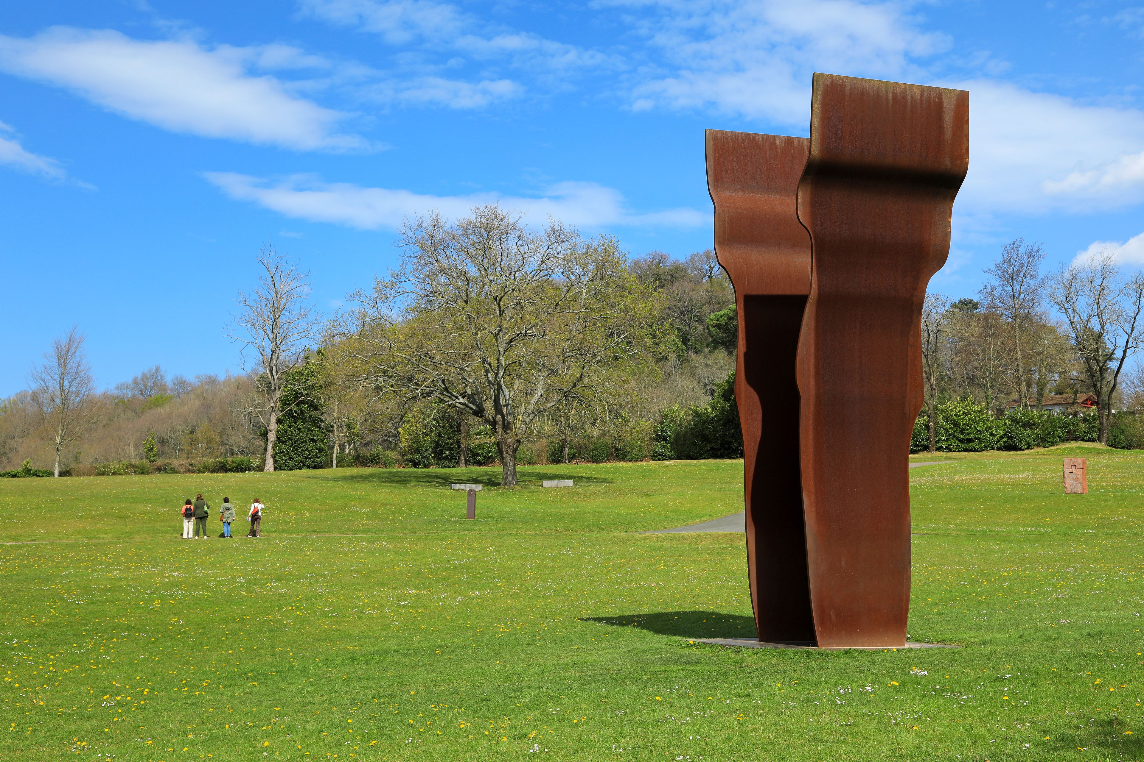Una escultura grande de color óxido en un parque cubierto de césped, con árboles y cielo despejado.