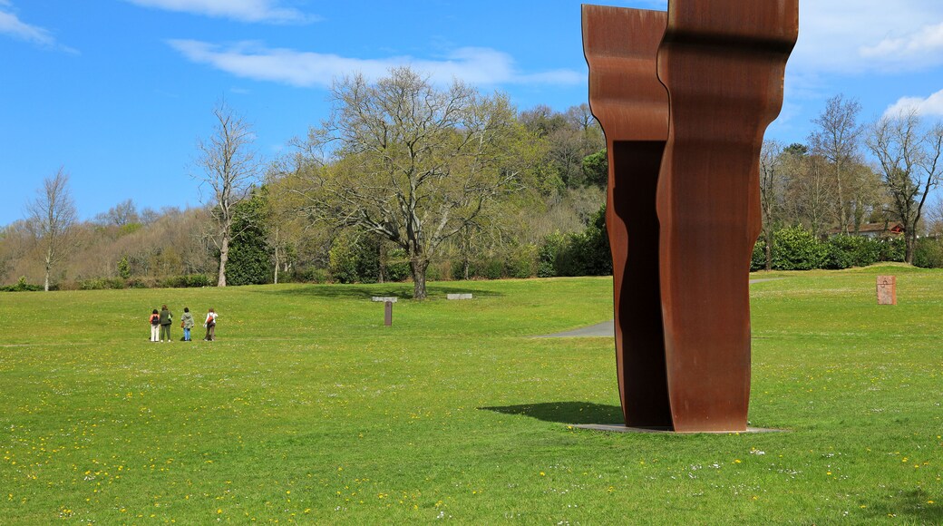 Una escultura grande de color óxido en un parque cubierto de césped, con árboles y cielo despejado.