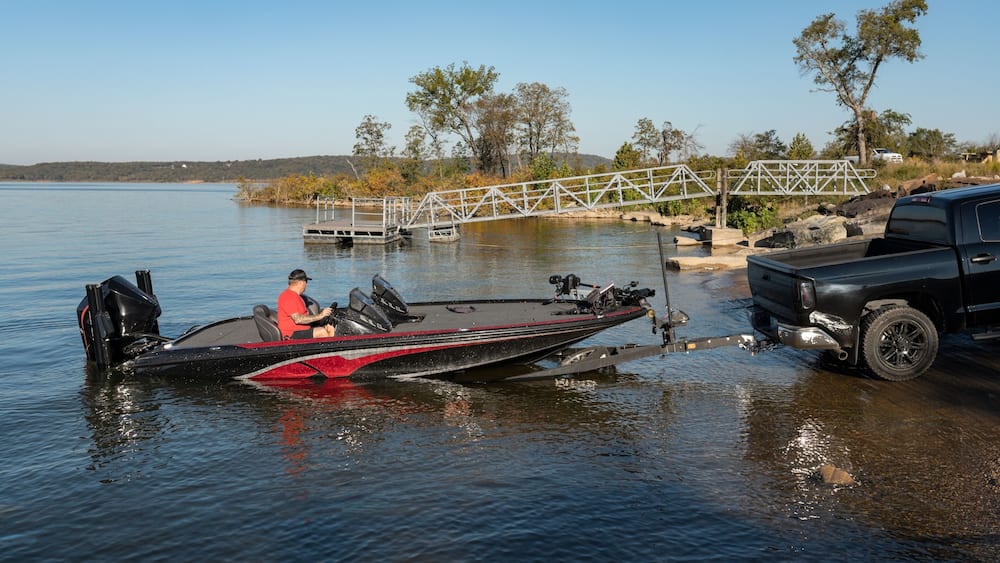 Un bateau noir avec une bande rouge, un homme portant une chemise rouge et un camion noir sur un lac calme, avec un pont métallique et des arbres en arrière-plan.