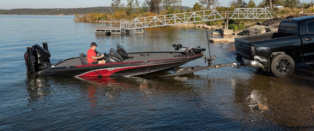 A black boat with a red stripe, a man in a red shirt, and a black pickup truck on a calm lake with a metal bridge and trees in the background.
