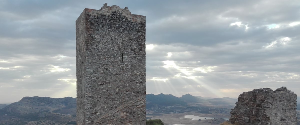 A tall, weathered stone tower amidst a mountainous landscape with a river in the distance.