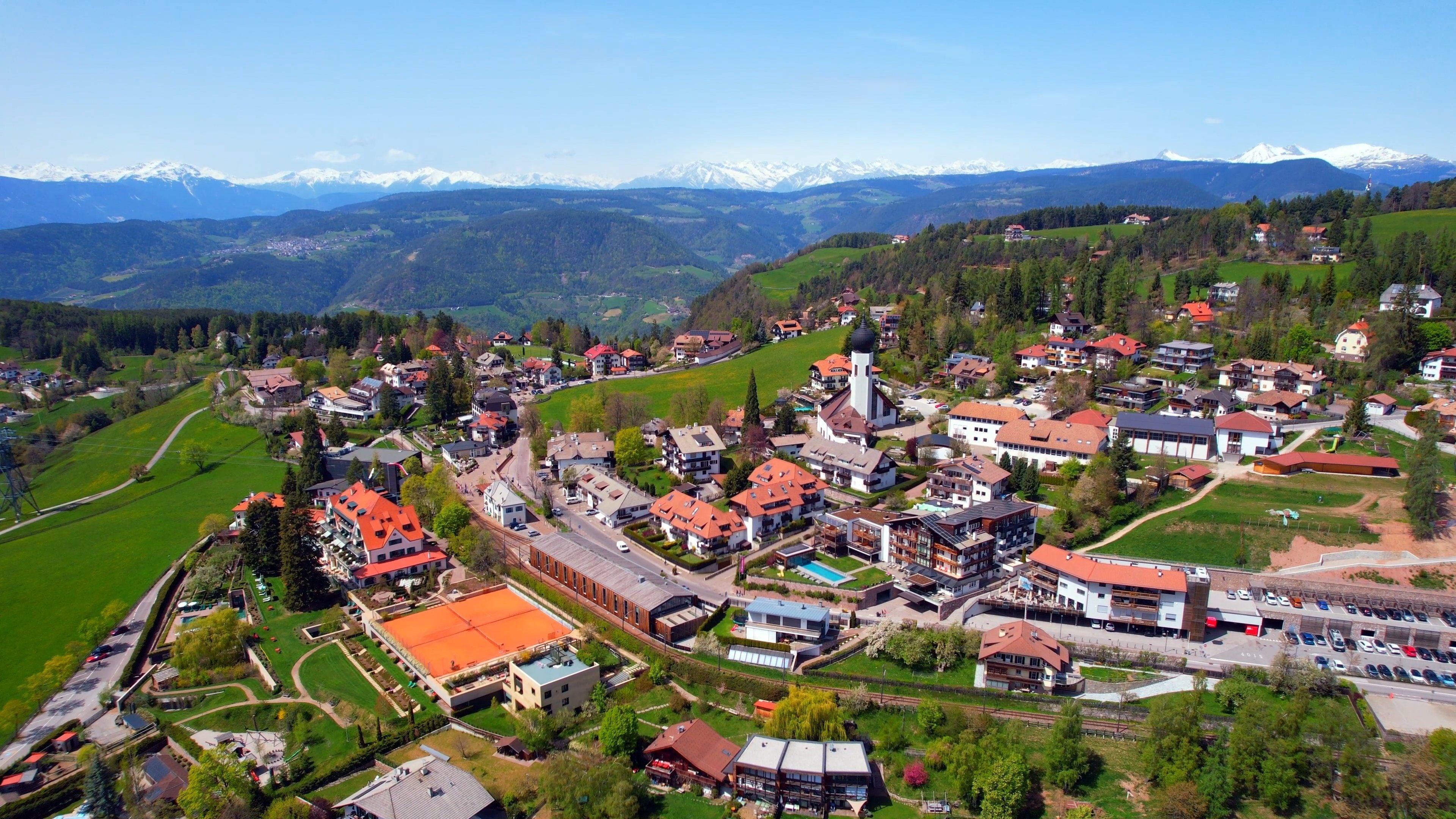 Un pueblo montañoso con picos nevados al fondo, rodeado de campos verdes exuberantes y edificios residenciales.