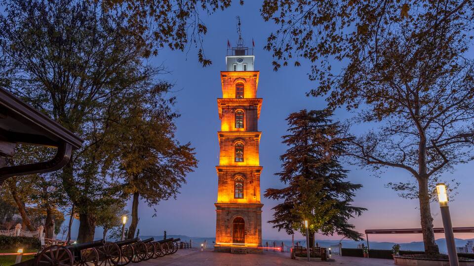 A lit tower with a clock, surrounded by trees and a paved walkway.