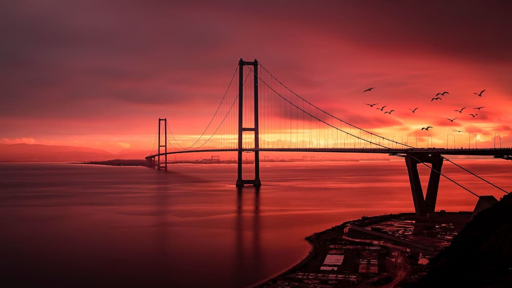 Eine Hängebrücke bei Sonnenuntergang mit ruhigem Wasser und einer Küstenlinie.