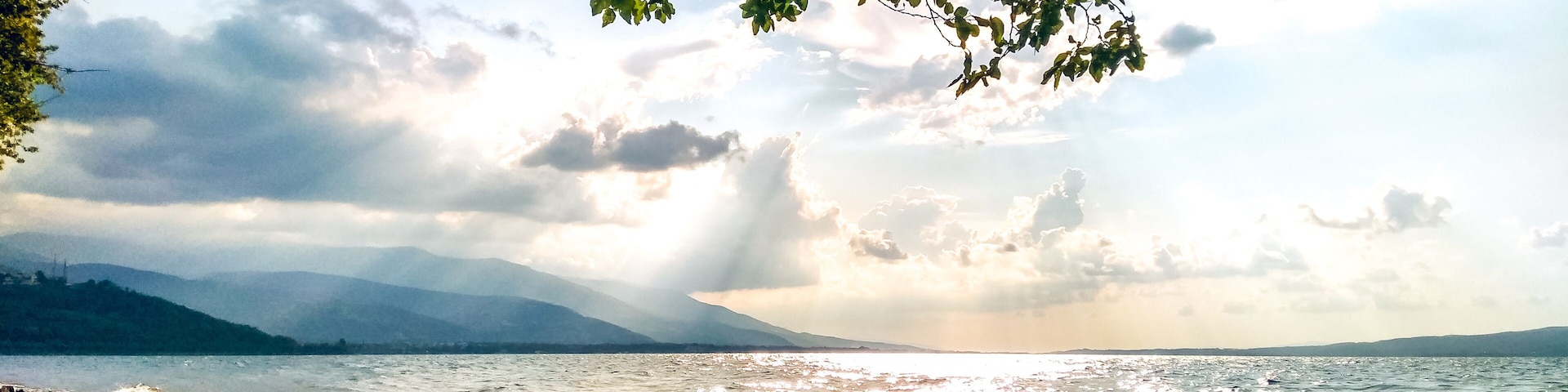 Uma costa rochosa com luz solar filtrando pelas árvores, um corpo d'água calmo e montanhas ao longe.