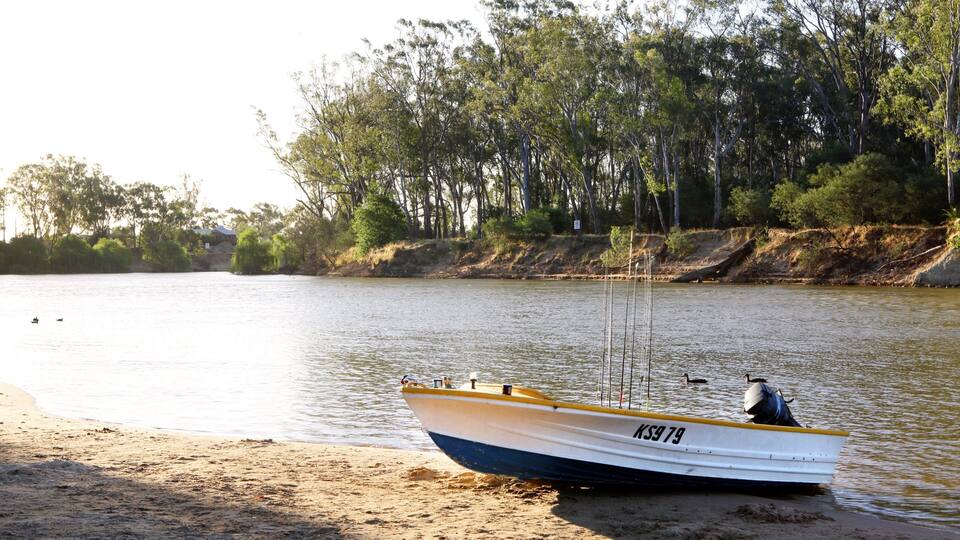 A small boat with the registration number KS979 on a sandy shore by a calm river, surrounded by dense trees.
