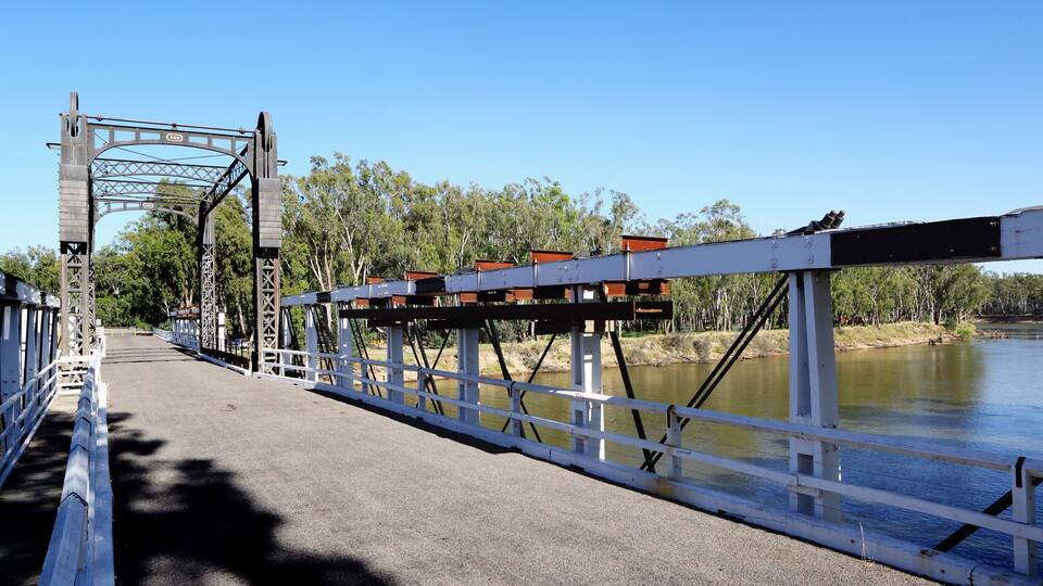 A bridge with a metal structure, a paved walkway, and a body of water with trees in the background.
