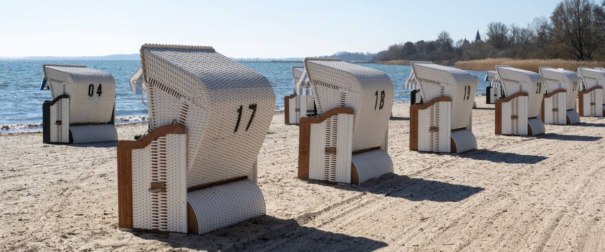 A row of beach chairs numbered 04 to 22 on a sandy beach with a body of water and trees in the background.