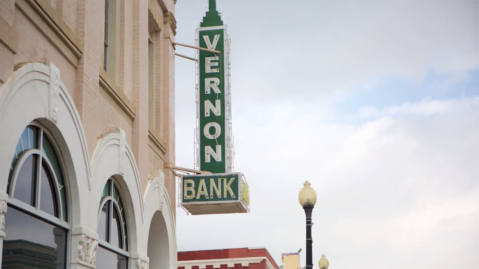 A vintage bank sign with the name 'VERNON BANK' in white letters on a green background, attached to a building with arched windows and a street lamp nearby.