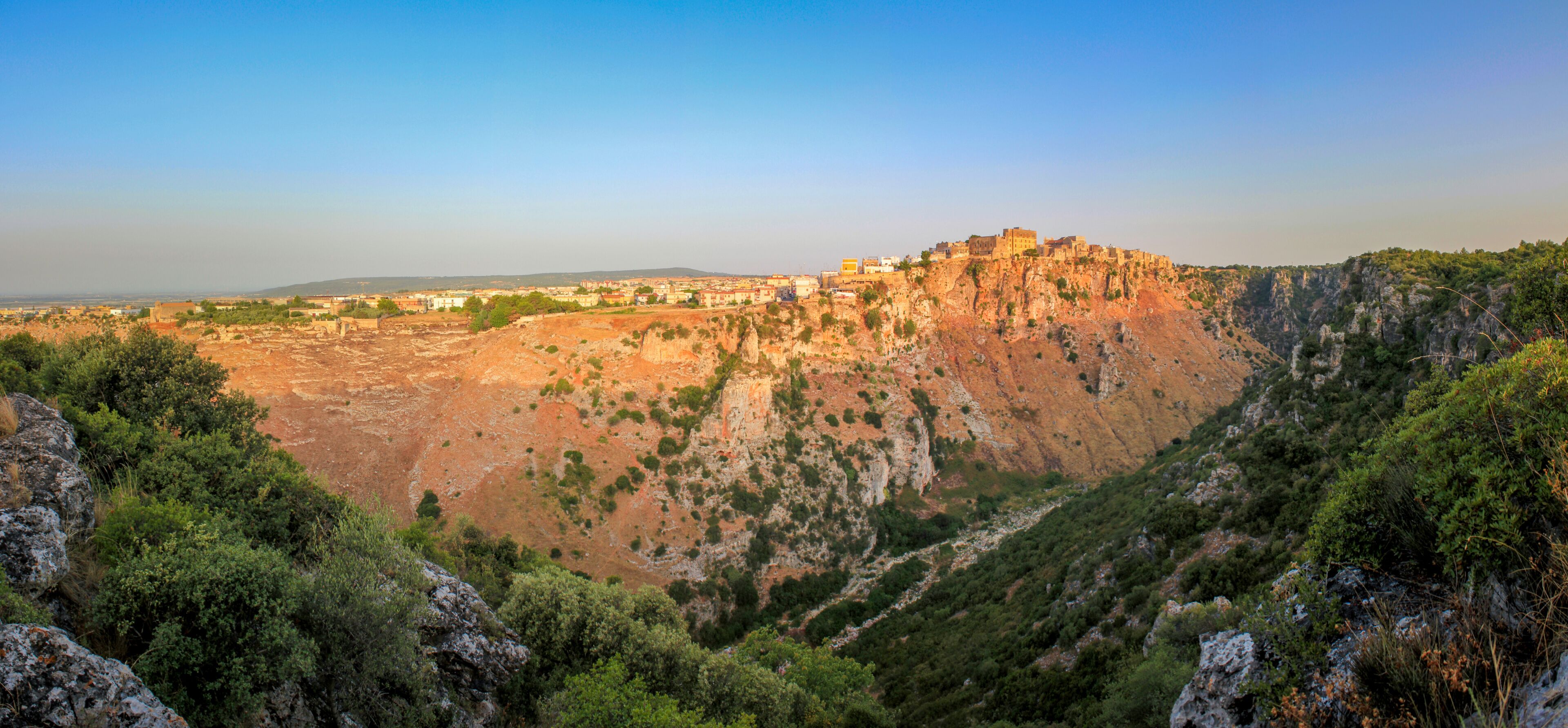 A rugged mountain with a fortified structure on top, surrounded by dense greenery and rocky terrain.
