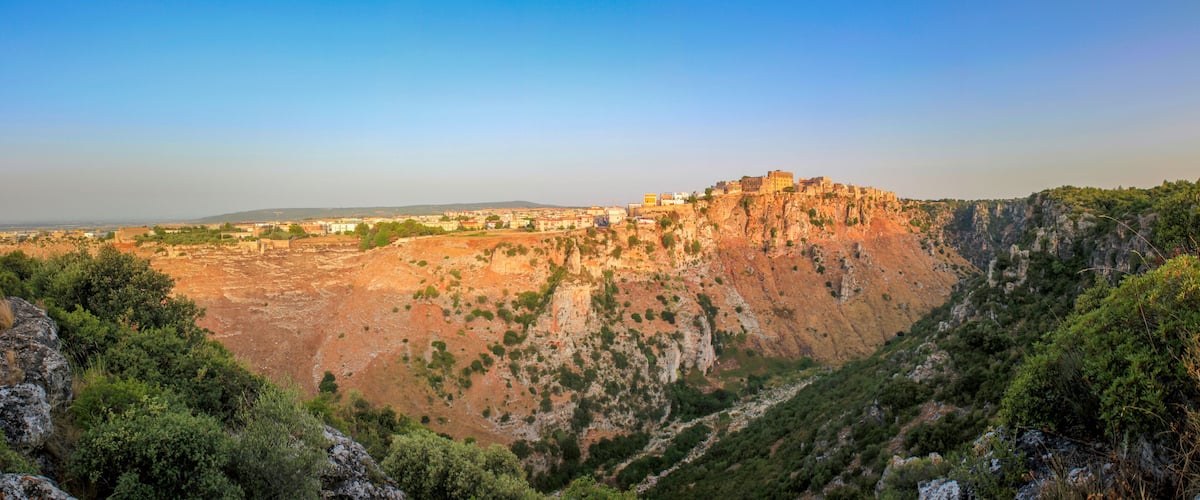 A rugged mountain with a fortified structure on top, surrounded by dense greenery and rocky terrain.