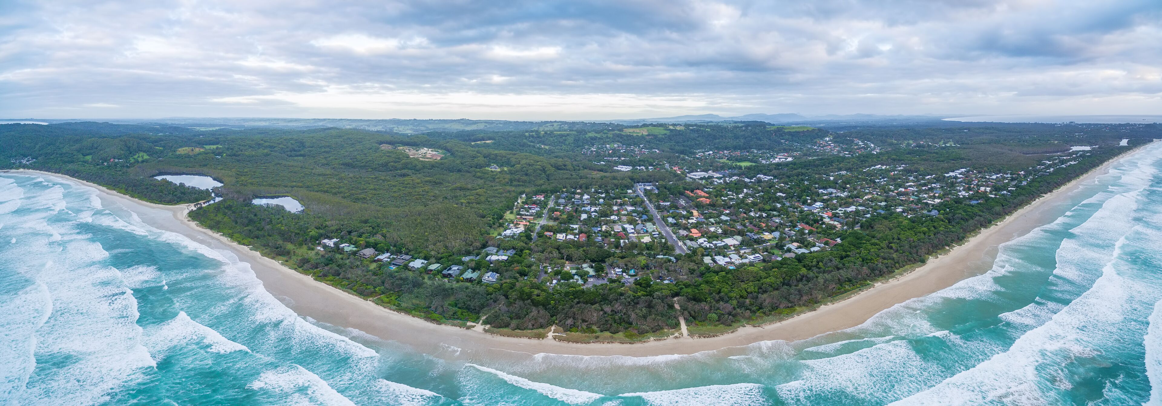 An aerial view of a coastal town with a beach, surrounded by dense forest, and a distant mountain range.