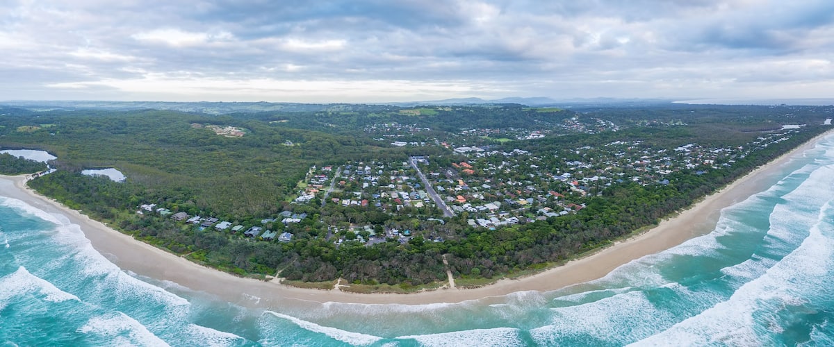 An aerial view of a coastal town with a beach, surrounded by dense forest, and a distant mountain range.