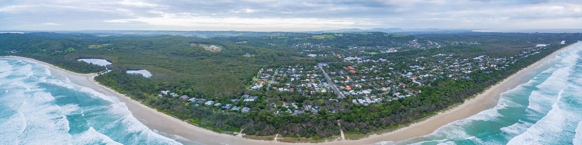 An aerial view of a coastal town with a beach, surrounded by dense forest, and a distant mountain range.