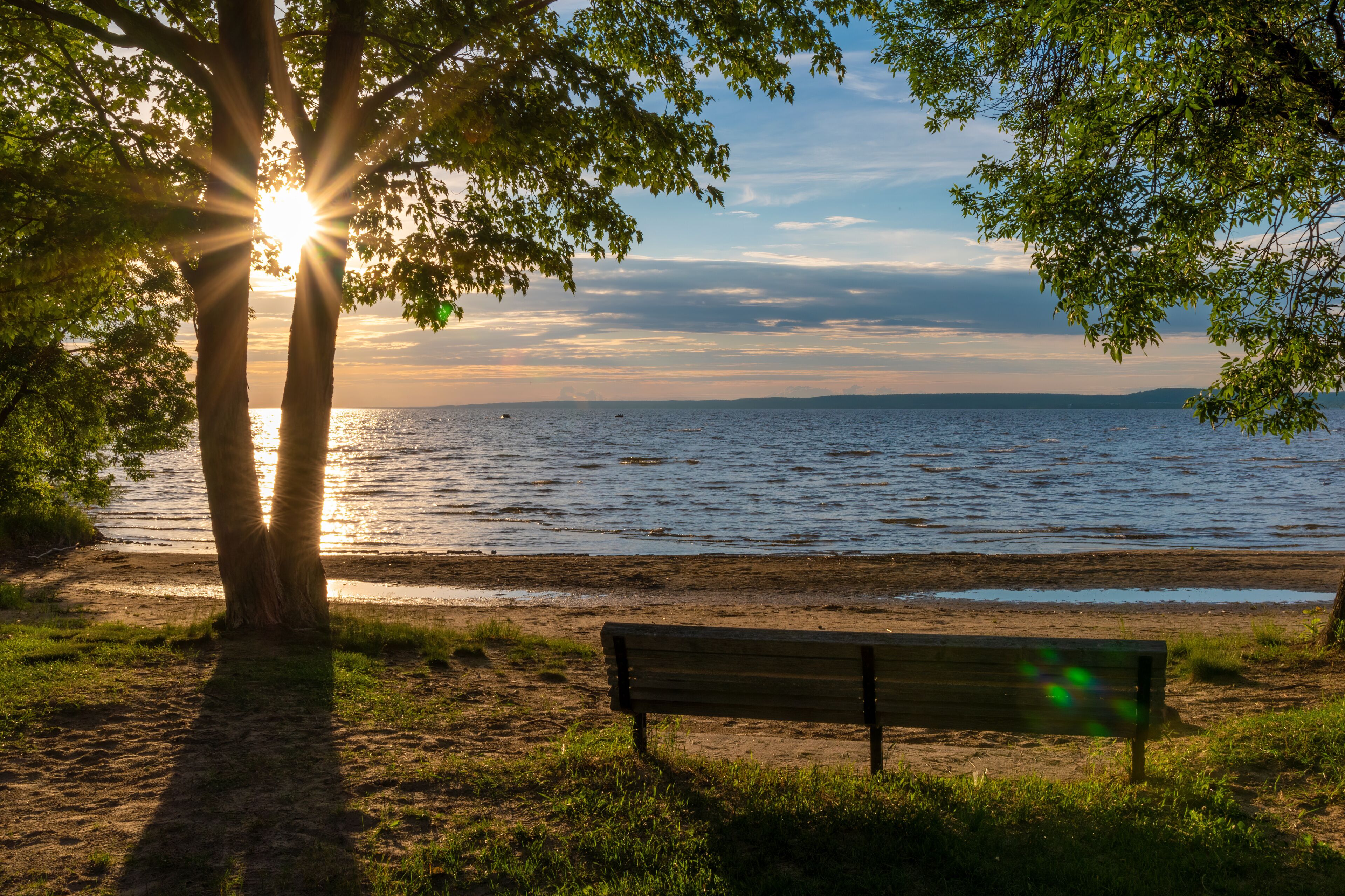 The sun pokes through a double tree trunk beside an empty park bench during a beautifully colourful sunset over Lake Nipissing in North Bay, Ontario.