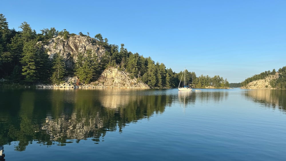 A calm lake with a sailboat, surrounded by dense forests and rocky cliffs.
