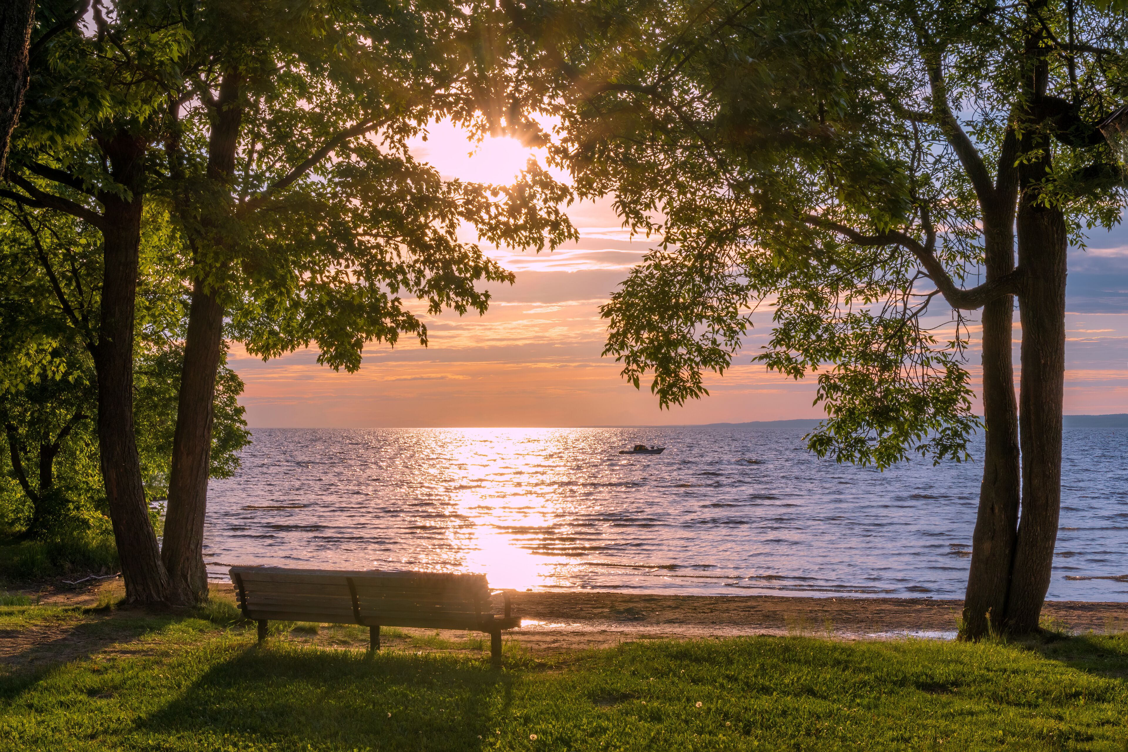 A pair of trees frames an empty park bench as a single boat floats on Lake Nipissing during a beautifully colourful sunset in North Bay, Ontario.