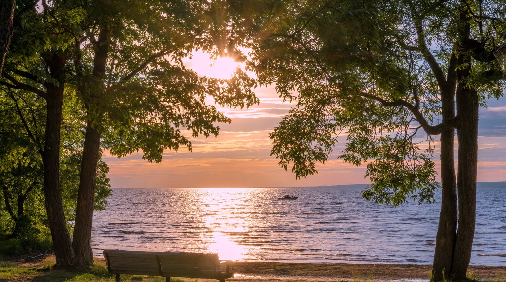 A pair of trees frames an empty park bench as a single boat floats on Lake Nipissing during a beautifully colourful sunset in North Bay, Ontario.