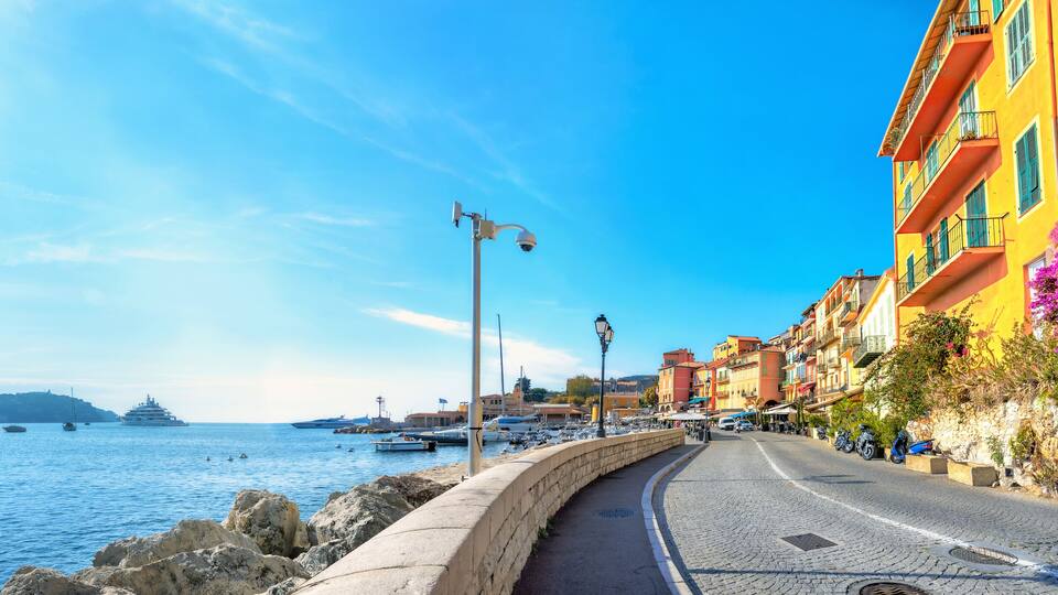 A coastal street with colorful buildings, a stone wall, and boats in the water.