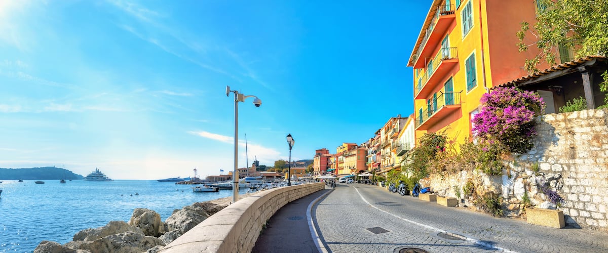 A coastal street with colorful buildings, a stone wall, and boats in the water.