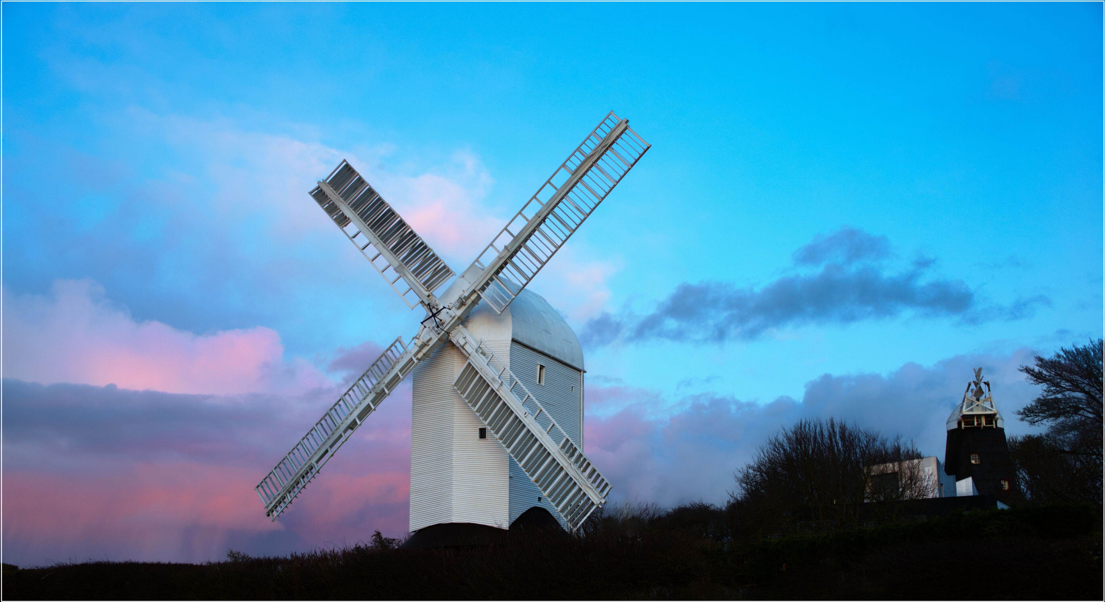 A windmill with a white body and a large cross-shaped sail, set against a twilight sky with pink and blue hues.