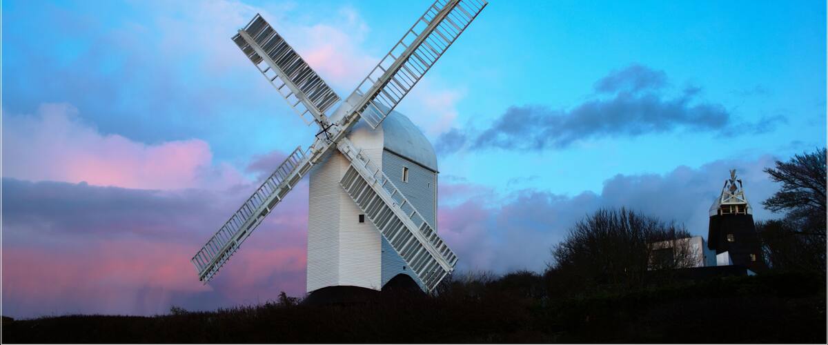 A windmill with a white body and a large cross-shaped sail, set against a twilight sky with pink and blue hues.