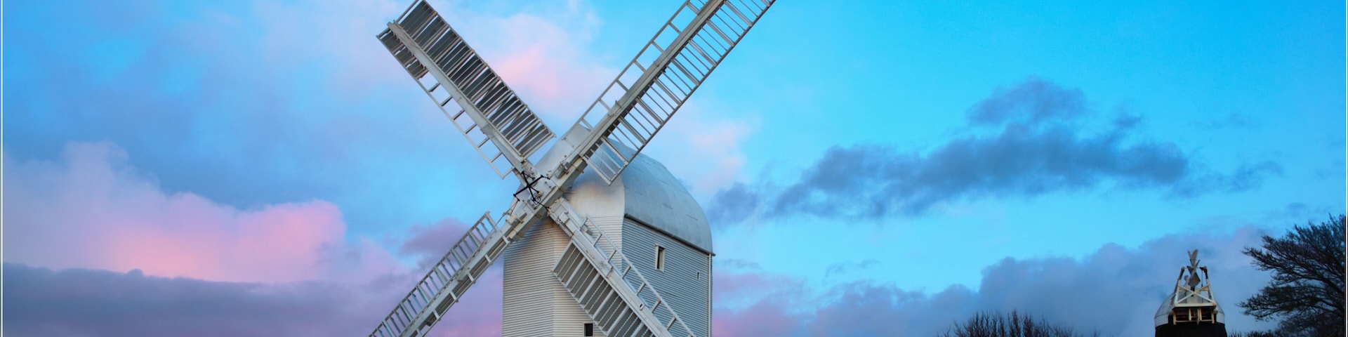 A windmill with a white body and a large cross-shaped sail, set against a twilight sky with pink and blue hues.