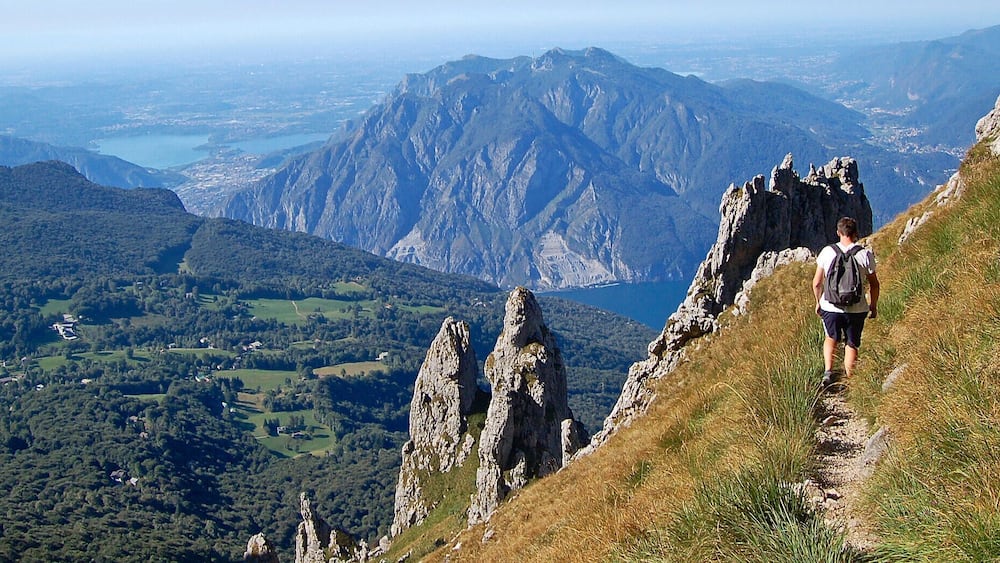 A hiker on a mountain trail with rocky outcrops, a valley with scattered houses, and a lake in the distance.