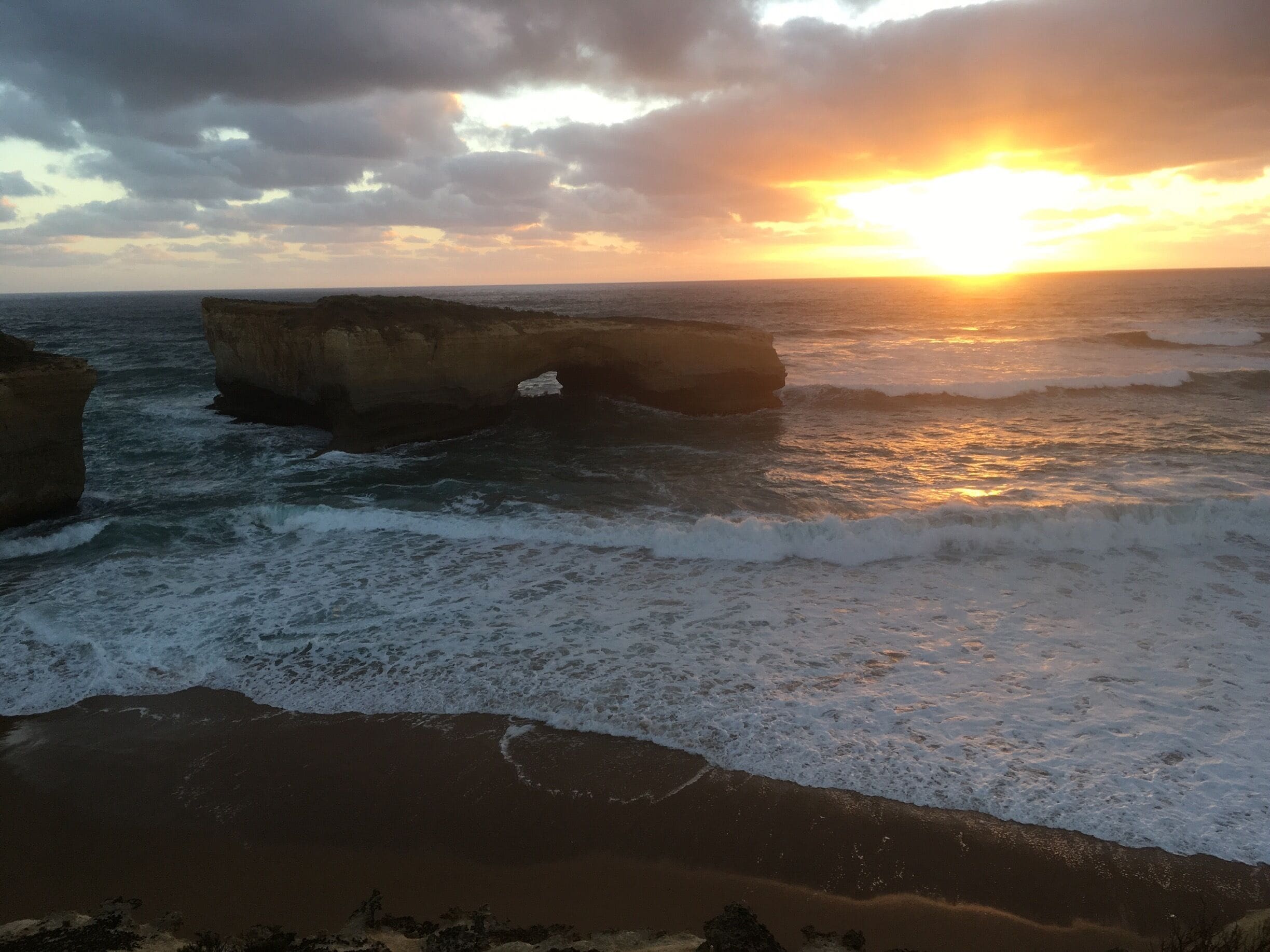 A sunset over a rocky coastline with waves crashing against large boulders.