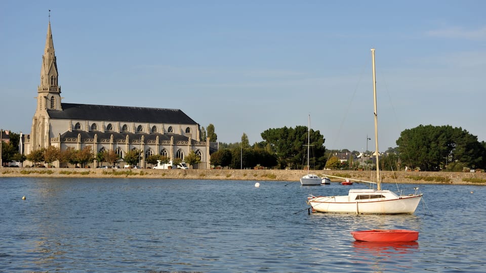 A church with a tall spire, a calm river, and boats.