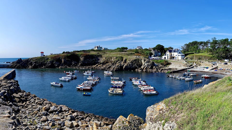 A coastal harbor with numerous boats docked, rocky shores, and buildings on the hillside.