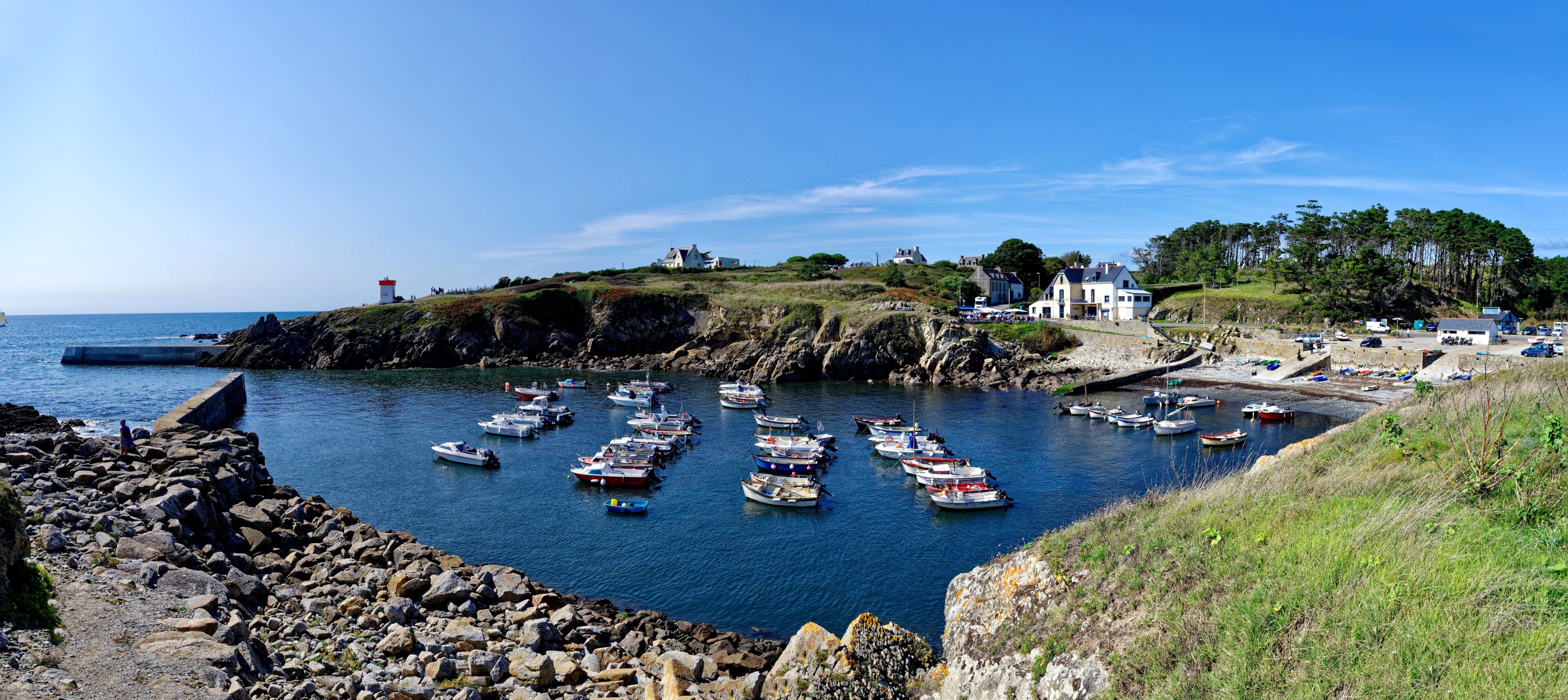 A coastal harbor with numerous boats docked, rocky shores, and buildings on the hillside.