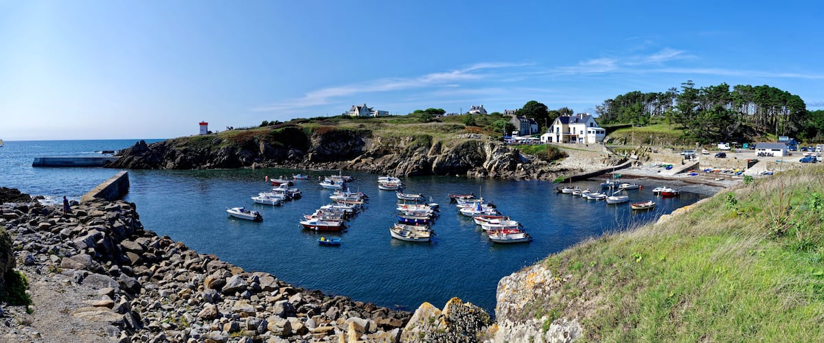 A coastal harbor with numerous boats docked, rocky shores, and buildings on the hillside.
