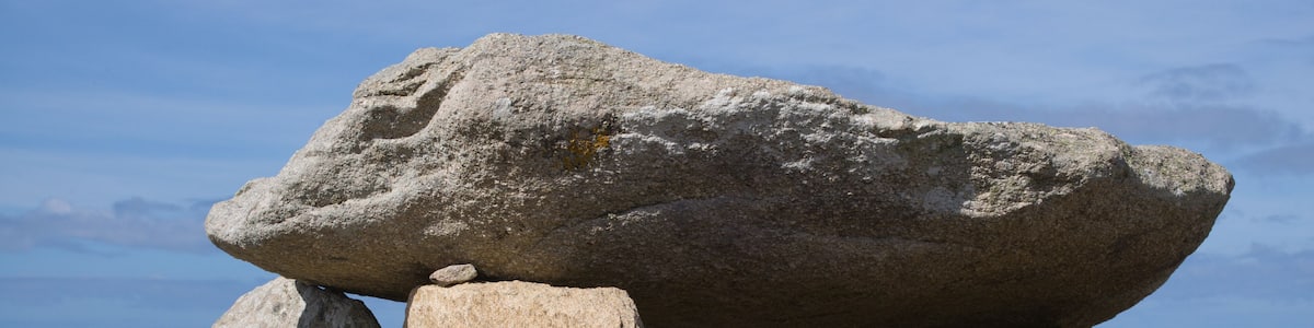 A large standing stone structure with a balanced stone on top, surrounded by other stones and the ocean in the background.