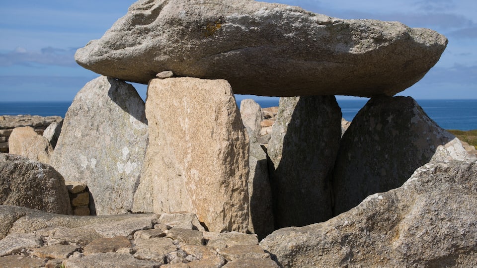 A large standing stone structure with a balanced stone on top, surrounded by other stones and the ocean in the background.