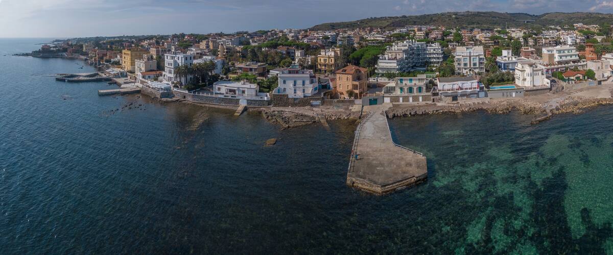 An aerial view of a coastal town with a mix of modern and traditional buildings, a clear blue sea, and a rocky breakwater.