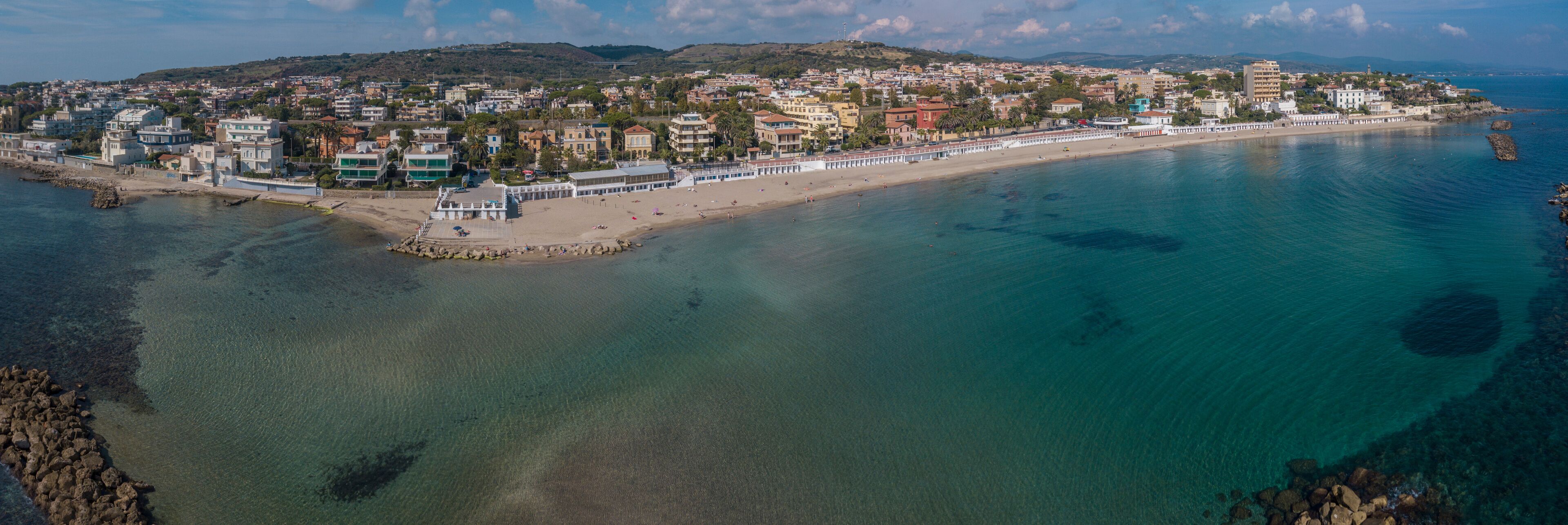 A coastal town with a beach, clear waters, and a hilly background.