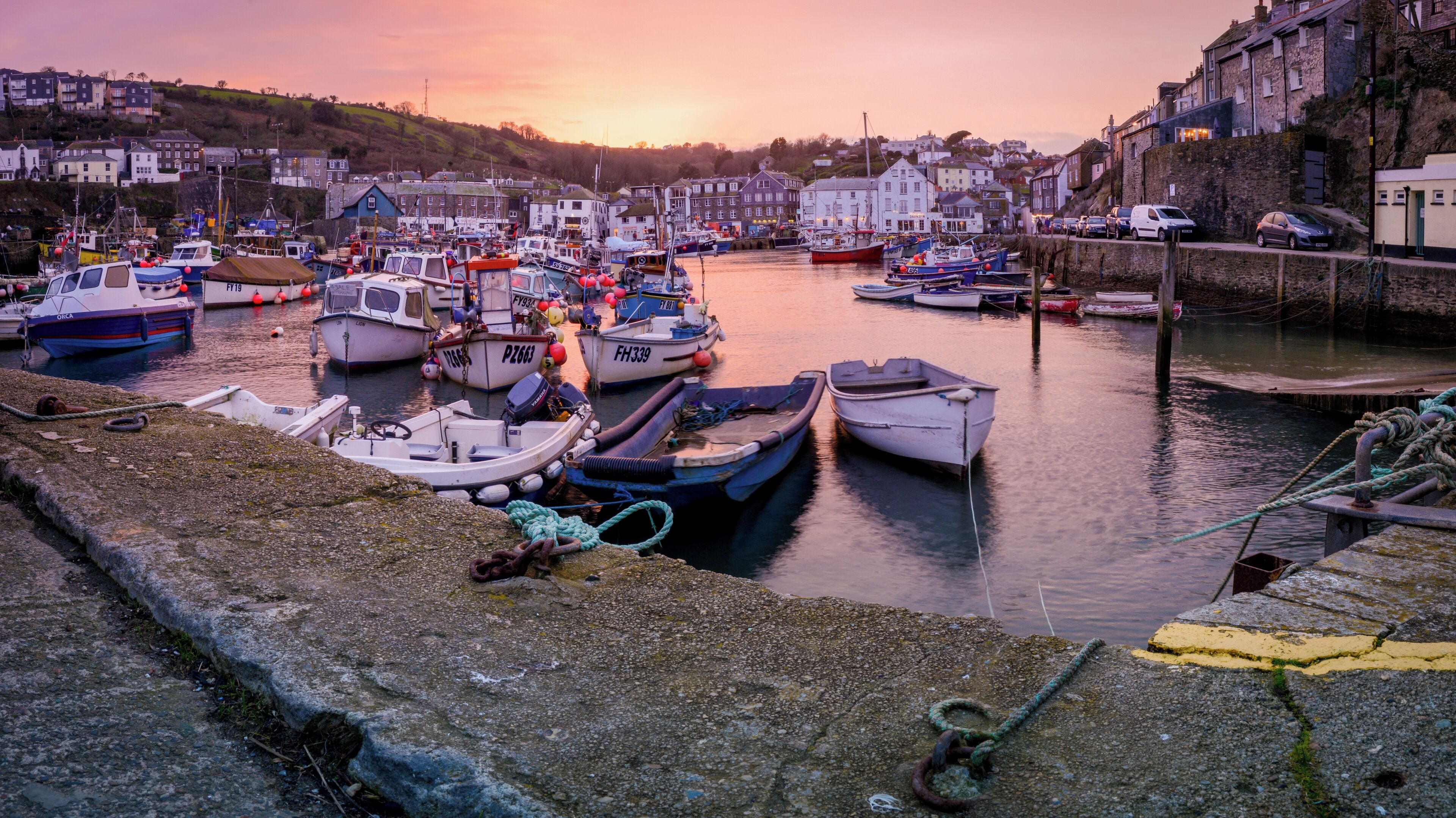 A harbor with various boats docked, surrounded by buildings and a sunset in the background.