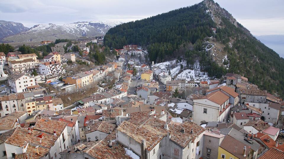 A town with red-tiled roofs nestled in a valley, surrounded by mountains with snow-capped peaks.