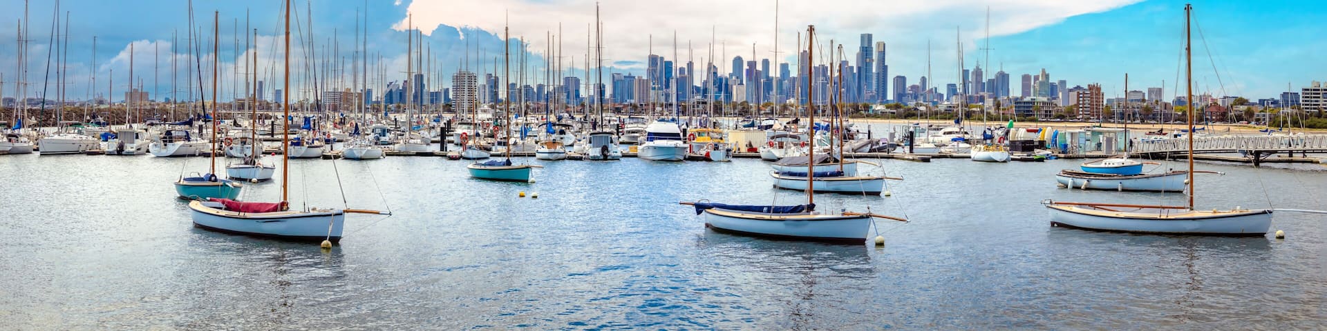 A marina with numerous sailboats docked, a city skyline in the background, and a partly cloudy sky.