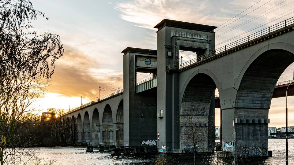 A concrete bridge with multiple arches over a river, with graffiti on the pillars and a sunset in the background.